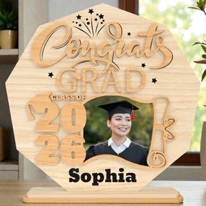 May include: Wooden graduation photo frame with the words "Congrats Grad" and "Class of 2026". The frame has a photo of a graduate and the name "Sophia" at the bottom. The frame is light brown and has a star and firework design.