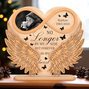 Puede incluir: Placa conmemorativa de madera en forma de corazón con alas de ángel. Presenta una foto, el nombre "Nathan" con fechas y el texto "Ya no a mi lado, sino para siempre en mi corazón". Mariposas negras decoran el diseño.