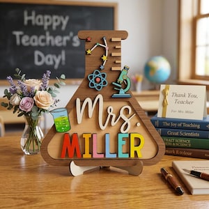 May include: A wooden teacher appreciation gift shaped like a science flask. It features the name "Mrs. Miller" in colorful letters, science-themed cutouts, and the text "Happy Teacher's Day!" on a chalkboard in the background.