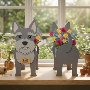 May include: Two gray Schnauzer-shaped planters, each holding colorful paper flowers. One planter faces forward, displaying a name tag that reads "Buddy." The other planter shows the dog's rear. The planters are on a wooden surface near a window.