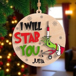 May include: Wooden Christmas ornament with the text "I WILL STAB YOU" in black, red, and green. The ornament features a green hand holding a syringe and a red stethoscope. The name "Julia" is at the bottom.