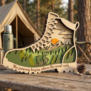 Puede incluir: Una pieza de arte de madera en capas con forma de bota de senderismo, que presenta un paisaje montañoso con árboles, un sol poniente y el texto "The journey begins where the road ends". La bota es de madera clara con detalles verdes y naranjas.