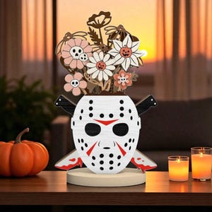 May include: A wooden Halloween-themed centerpiece featuring a white hockey mask with red accents and crossed knives. The mask holds wooden flowers with skull and pumpkin details. An orange pumpkin and lit candles sit nearby.