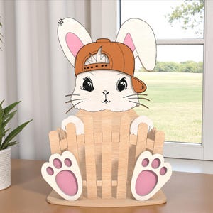 Peut inclure: Décoration de Pâques en bois représentant un lapin portant une casquette de baseball marron et tenant un panier. Le lapin a des oreilles blanches avec l'intérieur rose, un visage blanc et de grands yeux noirs. Le panier et les pieds sont en bois clair.