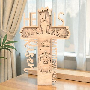 Puede incluir: Una cruz de madera con intrincados diseños recortados. Las palabras "HE IS RISEN" son visibles, junto con el texto "MATEO 28:6". La cruz presenta escenas de la crucifixión y la natividad, con estrellas y otros elementos decorativos.