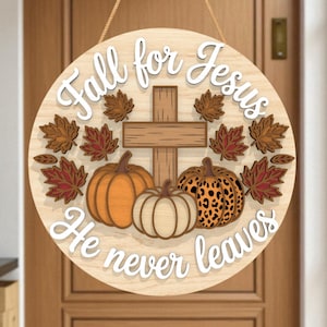 May include: Round wooden sign with the text "Fall for Jesus He never leaves". The sign features a cross, pumpkins, and fall leaves. The pumpkins are orange, white, and leopard print. The leaves are brown and red.