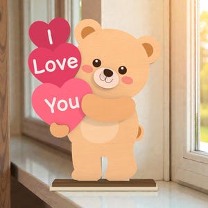 May include: A wooden bear figure holding three pink heart-shaped signs that read "I Love You". The bear has a light brown body, a cream-colored belly, and a small brown base. The background is a window with sunlight.