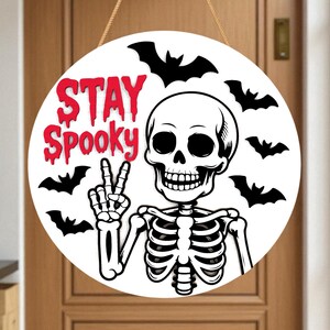 May include: Round white Halloween sign with a skeleton giving a peace sign and surrounded by black bats. The words "STAY Spooky" are in red, dripping font. The sign hangs from a brown rope.