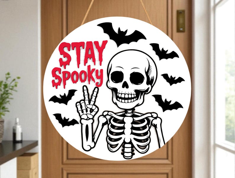 May include: Round white Halloween sign with a skeleton giving a peace sign and surrounded by black bats. The words "STAY Spooky" are in red, dripping font. The sign hangs from a brown rope.