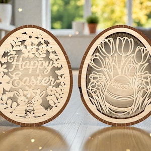 Puede incluir: Dos decoraciones de huevos de Pascua de madera. Un huevo presenta las palabras "Happy Easter" rodeadas de flores, conejitos y un huevo de Pascua. El otro huevo tiene un jarrón con tulipanes y narcisos. Ambos huevos tienen un borde marrón claro.