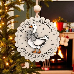 May include: A round, wooden Christmas ornament with a scalloped edge. The ornament features a cartoon goose wearing a bow tie, with the text "MERRY CHRISTMAS YOU SILLY GOOSE" around the edge. The ornament is hanging from a string.
