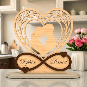 Può includere: Decorazione a forma di cuore in legno con la silhouette di una coppia che si bacia all'interno di una cornice a forma di cuore con un design ad albero. La base include un simbolo di infinito con i nomi "Sophia" e "Daniel" e dettagli a cuore.