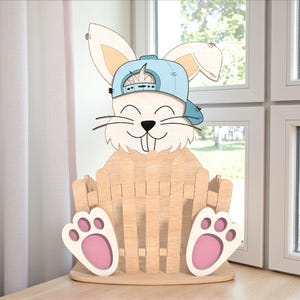 Peut inclure: Panier en forme de lapin en bois avec une casquette bleu clair. Le lapin a un visage souriant, de longues moustaches et de grandes oreilles. Le panier est de couleur marron clair avec des coussinets roses.