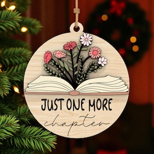 Puede incluir: Adorno navideño redondo de madera con diseño floral y texto "JUST ONE MORE chapter". El adorno presenta un libro abierto con flores y vegetación. Cuelga de una cuerda de hilo natural.