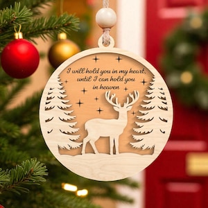 May include: Wooden Christmas ornament with a deer silhouette, trees, and the text "I will hold you in my heart, until I can hold you in heaven." The ornament is round and hangs from a string with a wooden bead.