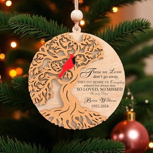 Op de afbeelding: Een houten kerstornament met een boomontwerp en een rode kardinaal. Het ornament heeft de tekst "Those we Love don't go away..." en de naam "Brian Williams 1952-2024". Het hangt aan een kerstboom.