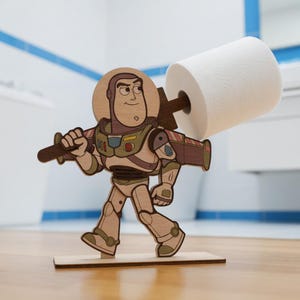 May include: A wooden Buzz Lightyear toilet roll holder. The holder is a laser-cut design of the character from the Toy Story film, holding a roll of white toilet paper. The holder is made of light brown wood with painted details.