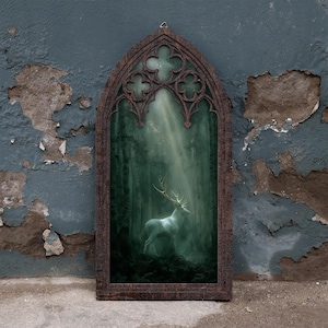 Può includere: Una decorazione da parete rettangolare con una cornice in legno marrone scuro e una parte superiore ad arco gotico a punta. Il pannello centrale mostra un bosco dai toni verdi con un cervo bianco tra gli alberi, illuminato da un raggio di luce.