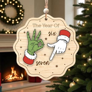 Op de afbeelding: Houten kerstornament met een uniek ontwerp. Het ornament toont twee handen, één groen en één wit, die de cijfers zes en zeven vormen. De tekst "The Year Of" staat bovenaan. Het ornament hangt voor een kerstboom.