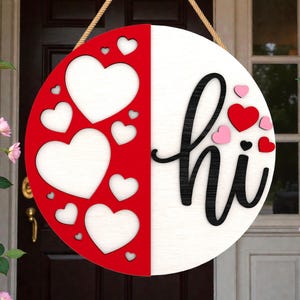 Puede incluir: Un letrero redondo de madera, mitad rojo con corazones recortados, mitad blanco con la palabra "hi" en escritura negra. Pequeños corazones rojos y rosas están esparcidos alrededor del texto. Una cuerda está adjunta para colgar.