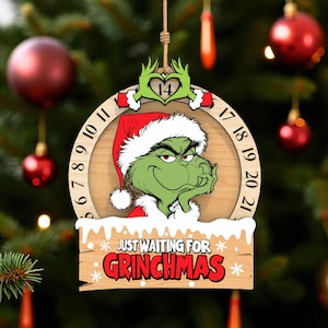 May include: Wooden Christmas ornament featuring the Grinch wearing a Santa hat. The ornament has a circular design with numbers and the text "JUST WAITING FOR GRINCHMAS". The Grinch is green with a grumpy expression. The ornament is hanging on a Christmas tree.