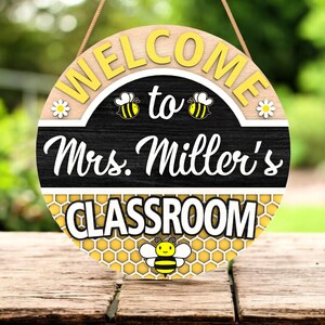 Peut inclure: Panneau rond en bois avec l'inscription "WELCOME to Mrs. Miller's CLASSROOM". Le panneau présente des lettres jaunes, des motifs d'abeilles et de marguerites, et un motif en nid d'abeille. Une ficelle marron est attachée pour l'accrocher.