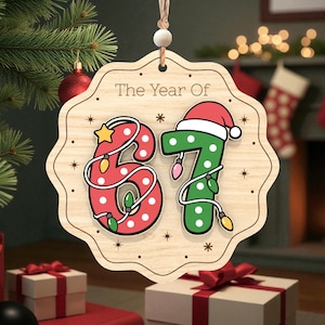 May include: Wooden Christmas ornament with the text "The Year Of" and the numbers "67". The number 6 is red with white polka dots and string lights. The number 7 is green with a Santa hat and string lights. The ornament is hanging from a string.