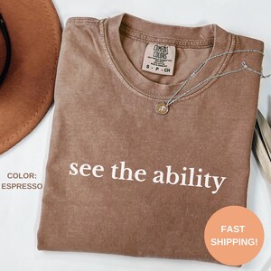 Puede incluir: Una camiseta marrón con el texto "see the ability" impreso en blanco. La camiseta está doblada y colocada sobre una superficie blanca.