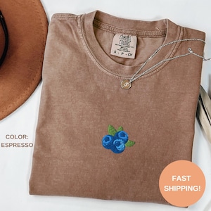 Op de afbeelding: Espresso-kleurig T-shirt met een klein geborduurd bosbessendesign. Het shirt heeft een ronde hals en een label met de tekst "Comfort Colors". Een zilveren ketting met een hanger ligt op het shirt. De tekst "COLOR: ESPRESSO" is zichtbaar.
