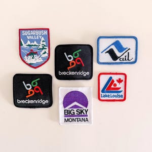 May include: A collection of embroidered patches featuring various ski resort logos. The patches are in different shapes and colors, including a Sugarbush Valley patch with a mountain scene, and a Big Sky Montana patch with a purple sun design.