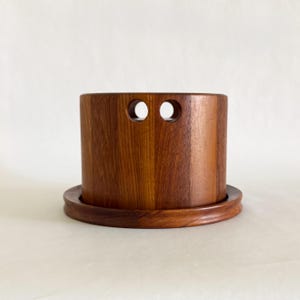 May include: A cylindrical wooden container with two circular holes near the top. The container sits on a matching round base. The wood grain is visible, and the color is a warm brown.