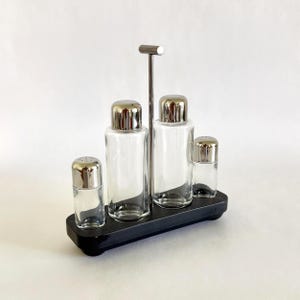 May include: A vintage salt and pepper set with a chrome handle and lids. The set includes two tall shakers, a small shaker, and a salt shaker, all in clear glass. The set is on a black tray.