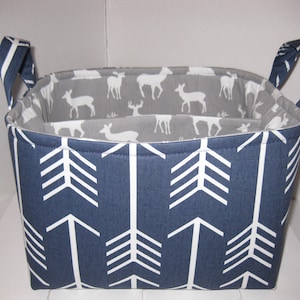 May include: A blue and white fabric storage basket with white deer silhouettes on a gray background and white arrow patterns on a blue background. The basket has two fabric handles.