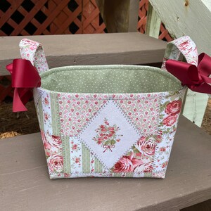 May include: A green and white fabric storage basket with red ribbon handles. The basket is decorated with a patchwork design of pink roses and green leaves. The basket is sitting on a wooden surface.
