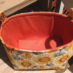 May include: A fabric basket with a sunflower pattern on the exterior and a red polka-dot interior. The basket has two handles and is designed for storage. The floral design features yellow sunflowers and green leaves on a light gray background.