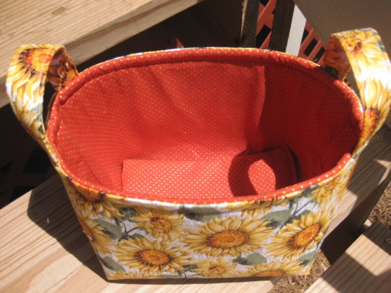 May include: A fabric basket with a sunflower pattern on the exterior and a red polka-dot interior. The basket has two handles and is designed for storage. The floral design features yellow sunflowers and green leaves on a light gray background.