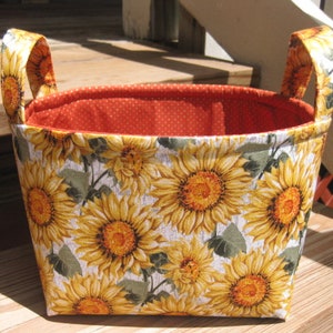 May include: A fabric storage basket with a vibrant sunflower pattern. The exterior features large yellow sunflowers with brown centers and green leaves on a white background. The interior is lined with orange fabric with white polka dots. The basket has two matching handles.