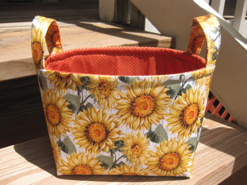 May include: A fabric storage basket with a vibrant sunflower pattern. The exterior features large yellow sunflowers with brown centers and green leaves on a white background. The interior is lined with orange fabric with white polka dots. The basket has two matching handles.