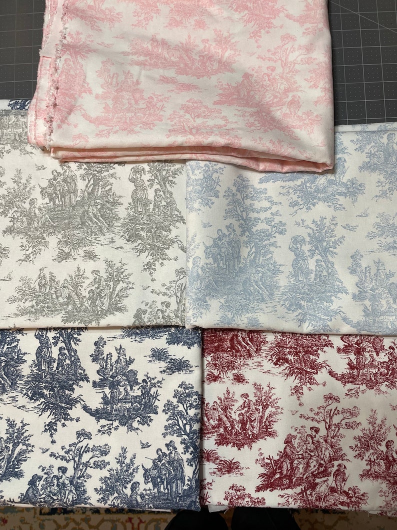 May include: Assortment of fabric pieces featuring a classic toile de Jouy pattern. The fabrics display scenes of figures and landscapes in various colors, including pink, gray, blue, navy, and burgundy, against a white background. The fabrics are folded and stacked.