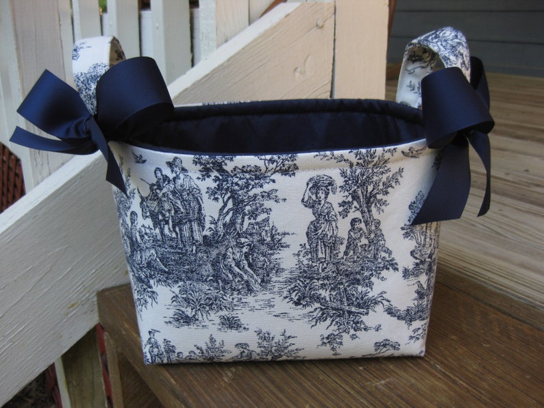 May include: A rectangular basket with a toile pattern in navy blue and white. The basket has navy blue bows on each side and navy blue fabric handles. The interior of the basket is navy blue.