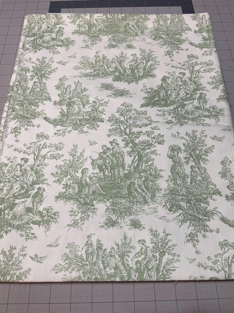 May include: A rectangular fabric with a white background and a green toile pattern. The pattern depicts scenes of people, trees, and birds. The fabric is likely used for home decor or crafting projects.