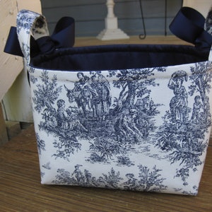 May include: A rectangular fabric basket with a toile pattern in navy blue and white. The basket has navy blue ribbon handles and a navy blue interior. The toile pattern depicts figures in a pastoral scene.