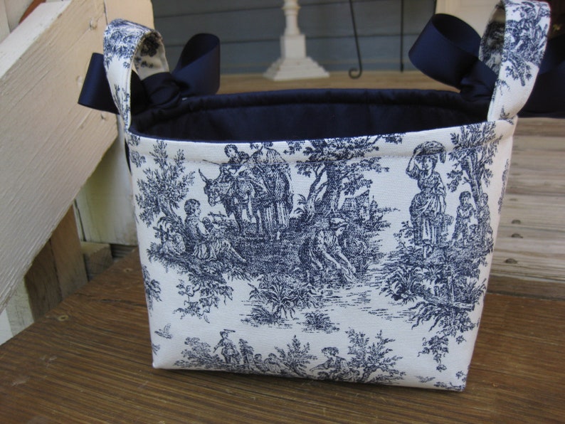 May include: A rectangular fabric basket with a toile pattern in navy blue and white. The basket has navy blue ribbon handles and a navy blue interior. The toile pattern depicts figures in a pastoral scene.