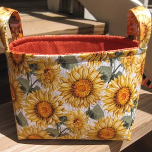 May include: A rectangular fabric basket with two handles. The exterior features a sunflower pattern with yellow and orange blooms on a white background. The interior is orange with white polka dots.