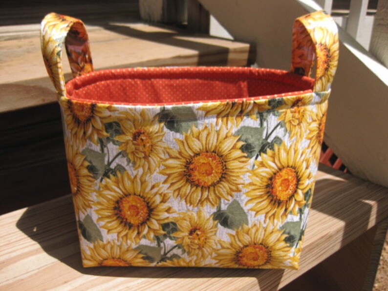 May include: A rectangular fabric basket with two handles. The exterior features a sunflower pattern with yellow and orange blooms on a white background. The interior is orange with white polka dots.