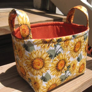 May include: A rectangular fabric basket with two handles. The exterior features a sunflower pattern with yellow and orange blooms on a white background. The interior is orange with white polka dots.