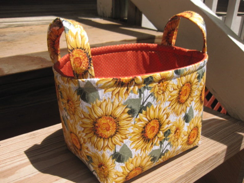 May include: A rectangular fabric basket with two handles. The exterior features a sunflower pattern with yellow and orange blooms on a white background. The interior is orange with white polka dots.