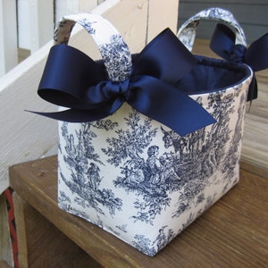 May include: A fabric basket with a toile pattern in blue and white. The basket has two handles and is adorned with a large navy blue bow on each side. The interior of the basket is navy blue.