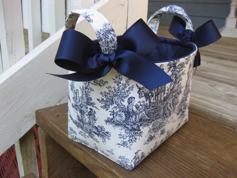 May include: A fabric basket with a toile pattern in blue and white. The basket has two handles and is adorned with a large navy blue bow on each side. The interior of the basket is navy blue.