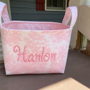 May include: A pink and white fabric basket with handles. The basket features a pink toile pattern and the name "Hanlon" embroidered in pink script. The interior is lined with solid pink fabric.